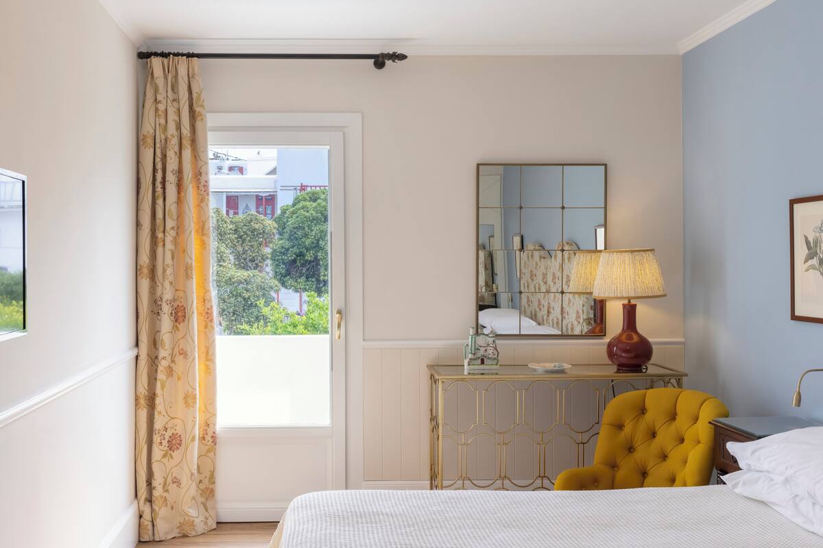 A hotel room with a bed, a yellow armchair, a bedside table with a lamp, a mirror, and a view of greenery outside.