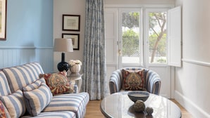A room with a striped sofa, two armchairs, a round table with a bowl, a lamp, and a window with curtains. - Kouneni Apartments (Mykonos)