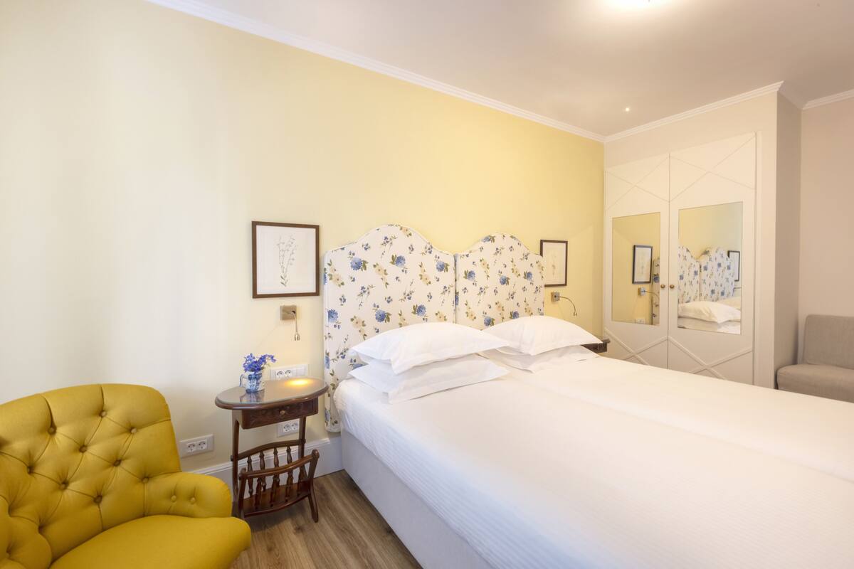 A bedroom with a bed, a yellow armchair, a small table with a vase, and a mirror.