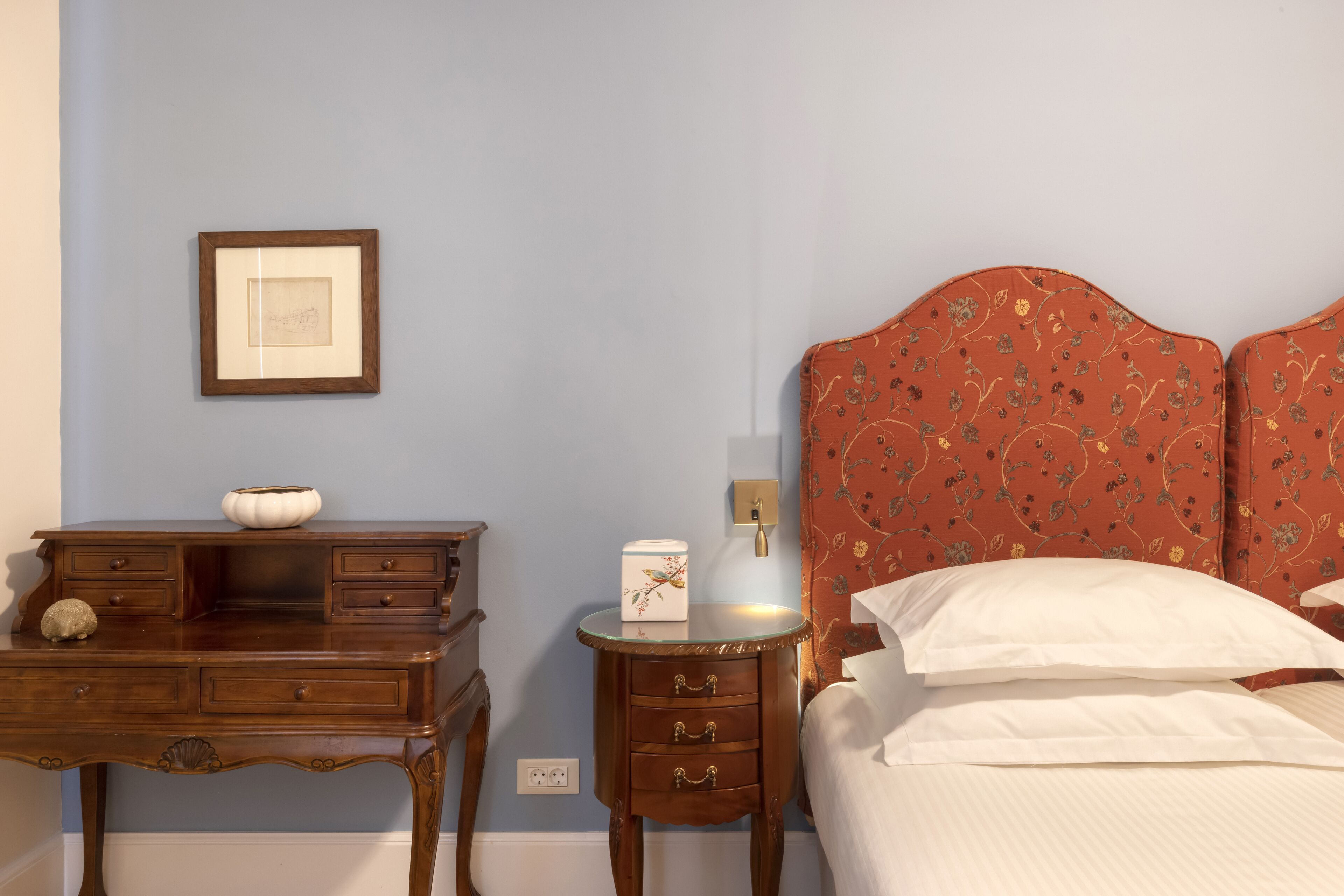 Un dormitorio con mesita de noche de madera, un cuadro enmarcado en la pared, una cama con cabecera estampada con motivos florales y una mesita de noche con lámpara.
