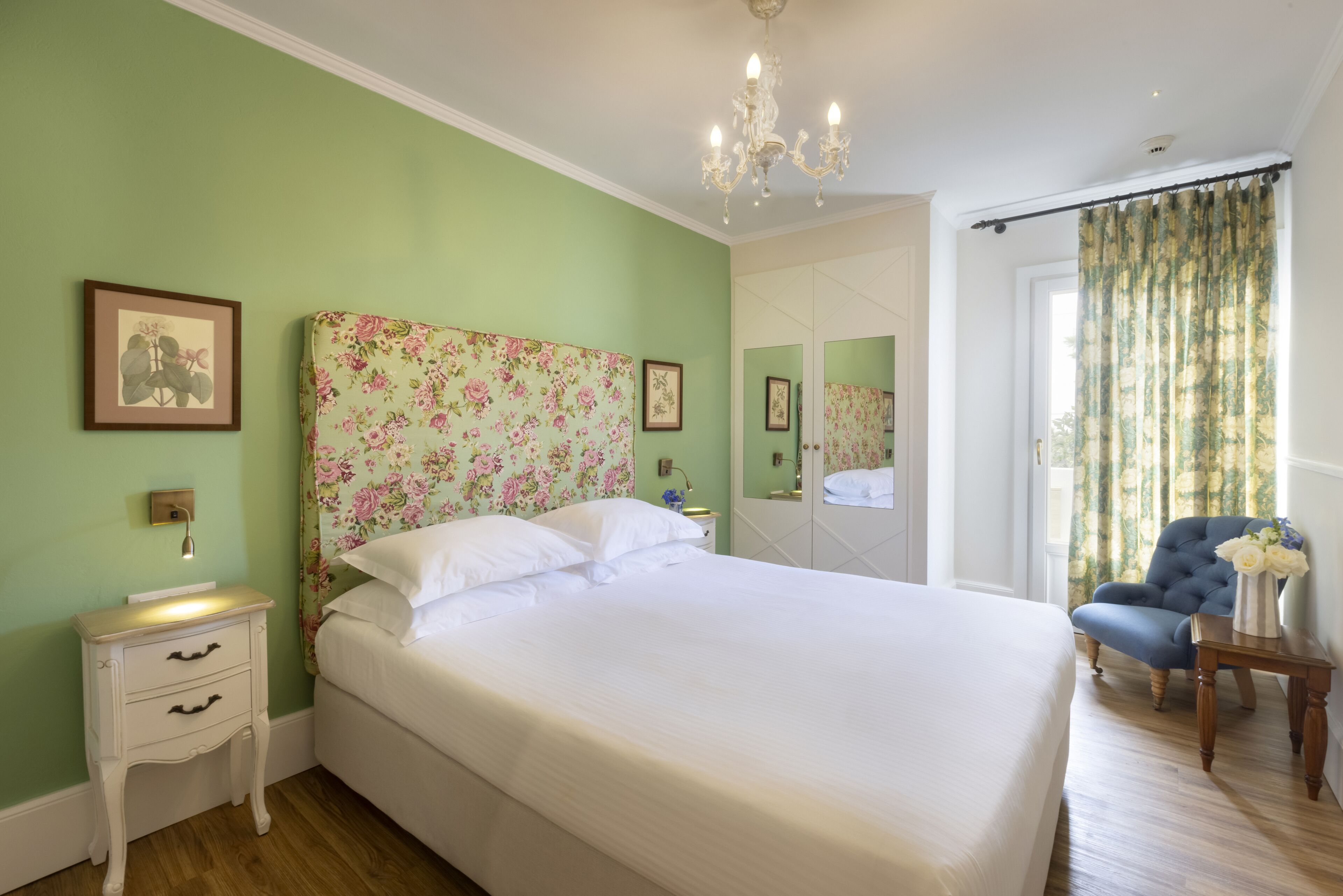 Un dormitorio con una cabecera floral, una mesita de noche con lámpara, una silla y un espejo grande.