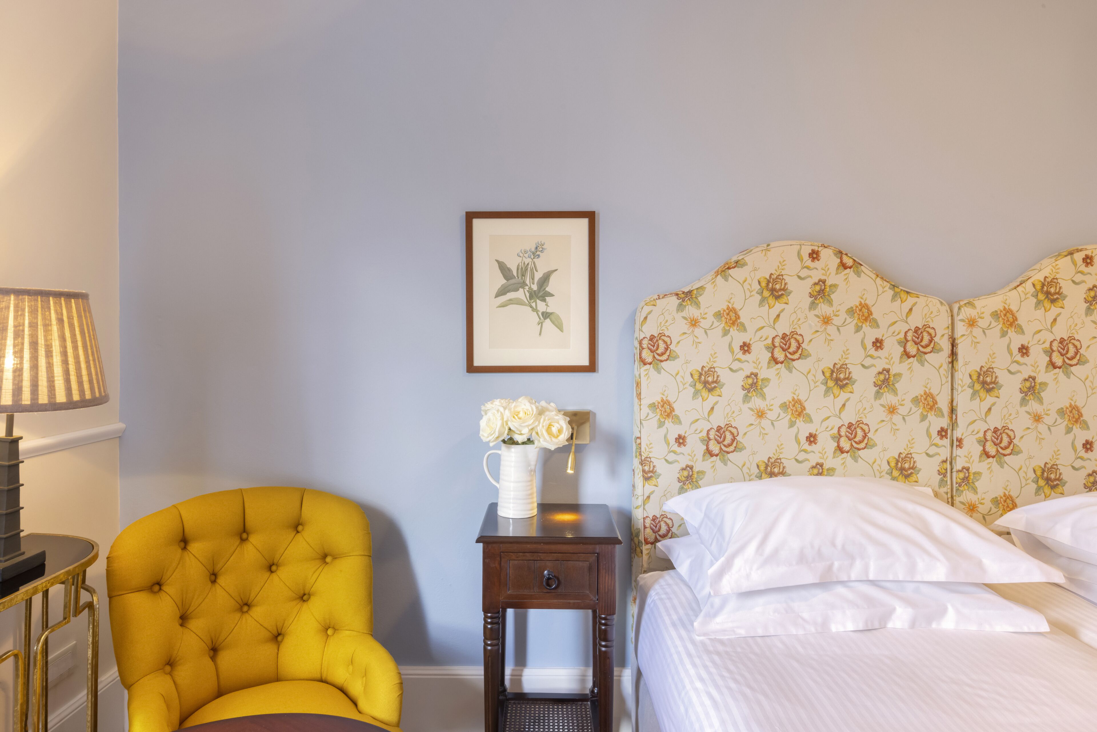 A bedroom with a floral headboard, a bedside table with a vase of flowers, a yellow armchair, and a lamp.