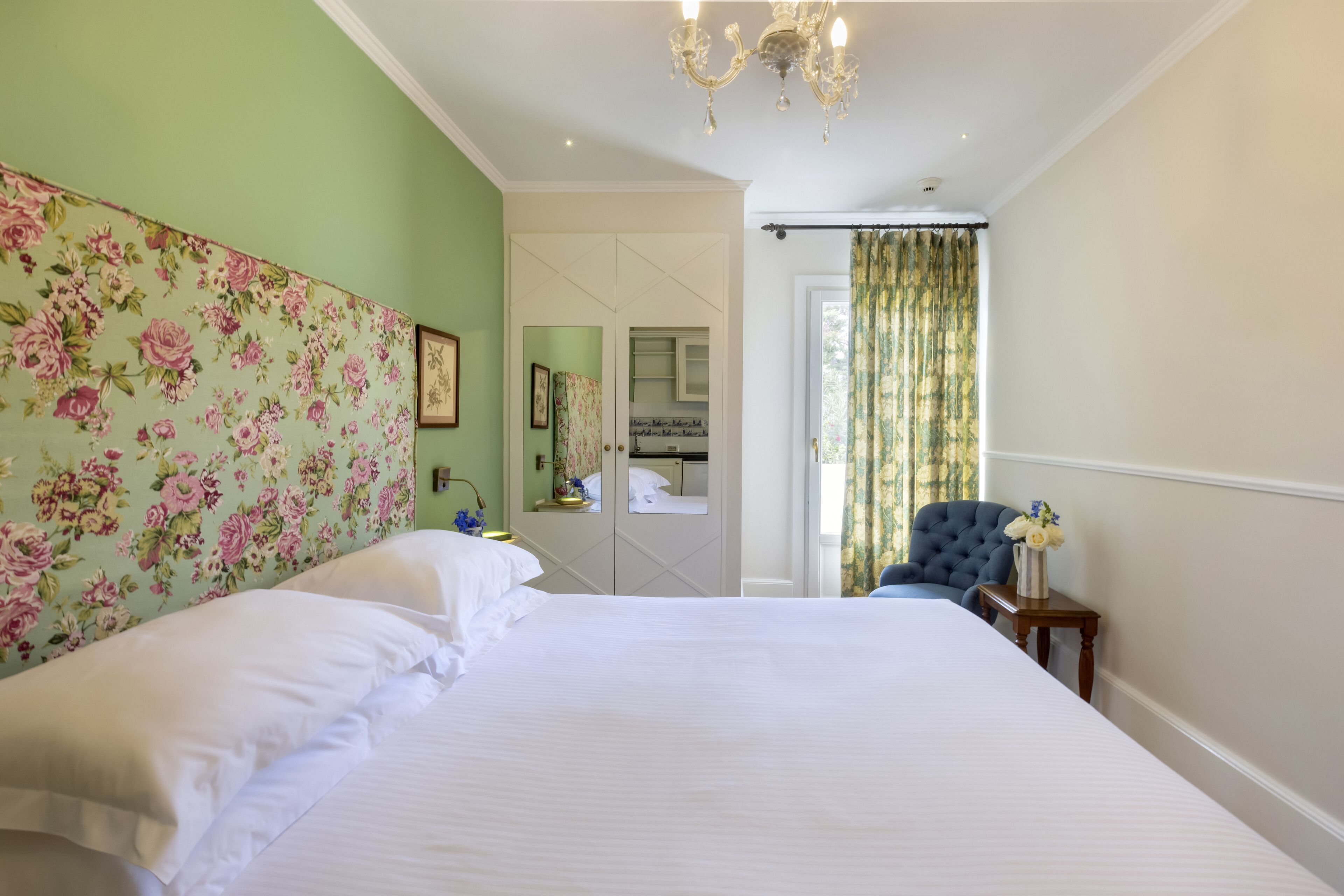 Una habitación de hotel con una cama grande, un cabecero floral, un espejo, un sillón azul y una mesita con un jarrón.