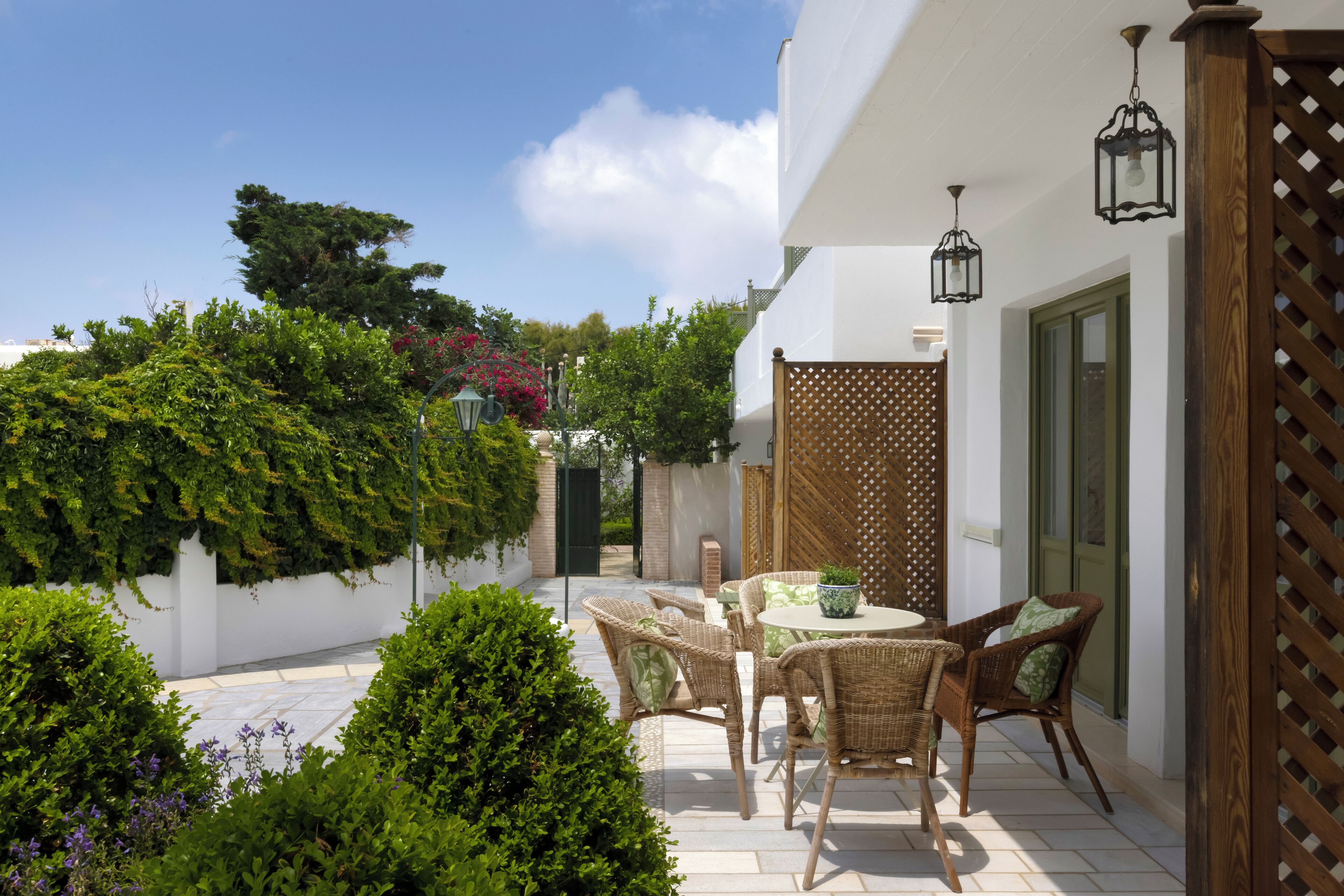 A patio with wicker furniture and a table, surrounded by greenery and a wooden fence.