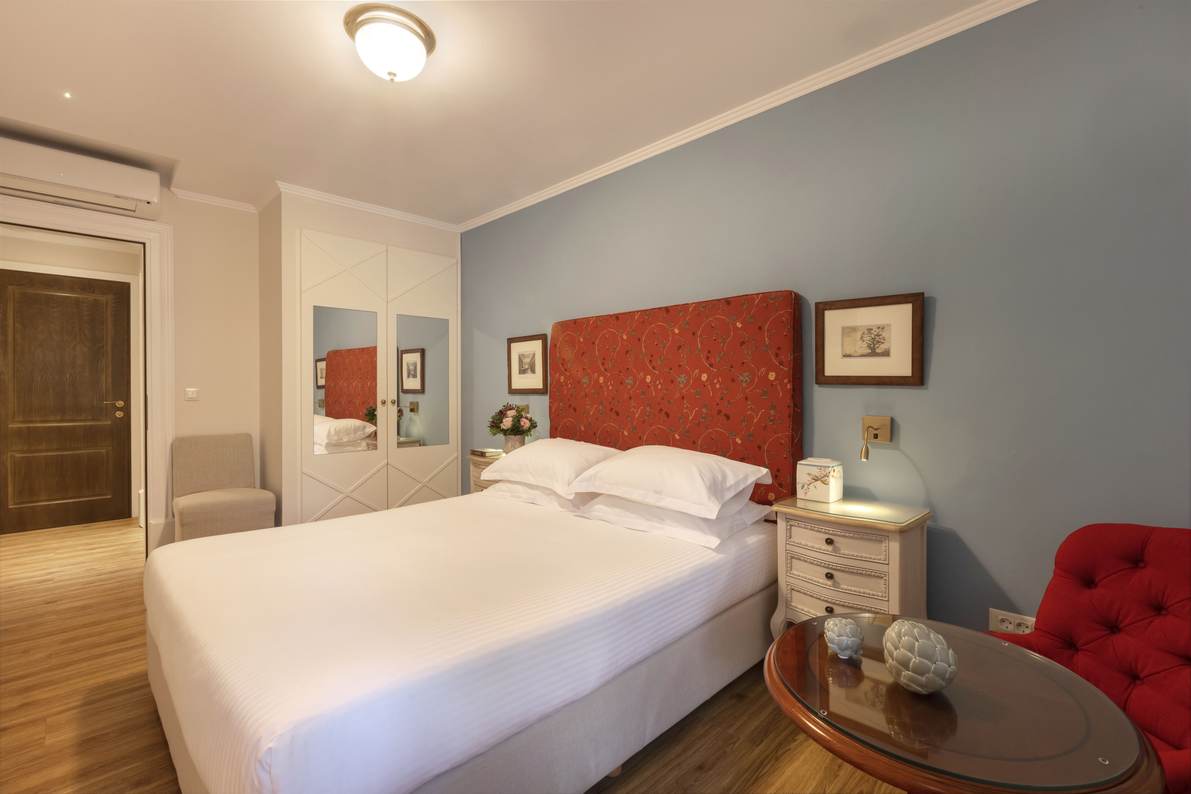 A neatly arranged hotel room with a large bed, a red upholstered chair, a small round table with decorative items, and a nightstand with a lamp.