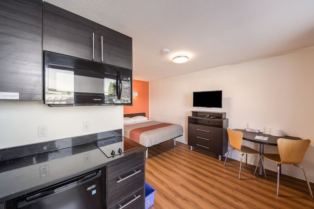 Una habitación de hotel con cama, escritorio con silla, televisión, microondas y horno empotrado.
