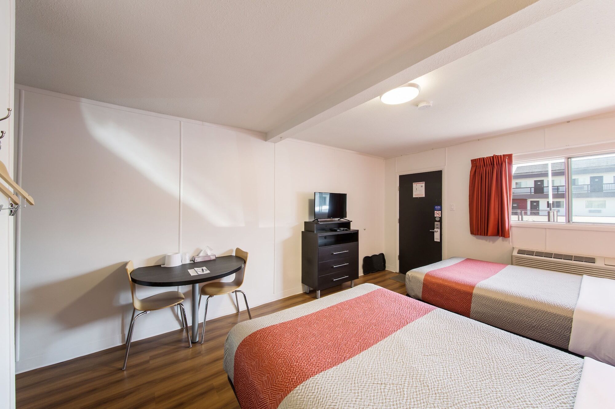 Habitación de hotel con dos camas, un escritorio y sillas, un televisor en una cómoda y una ventana con cortinas.