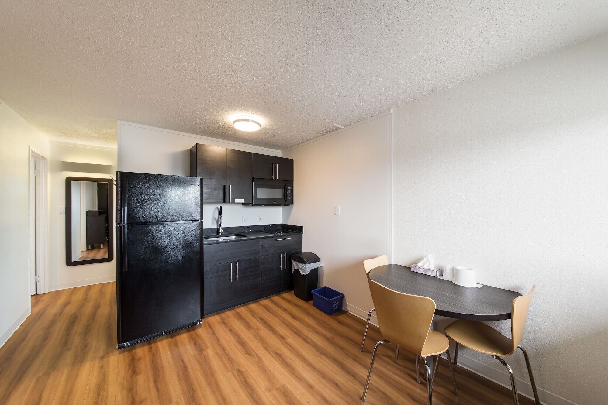Una cocina compacta con armarios oscuros, un refrigerador negro, un microondas y un lavamanos. Hay una mesa de comedor con dos sillas y un cubo de basura.