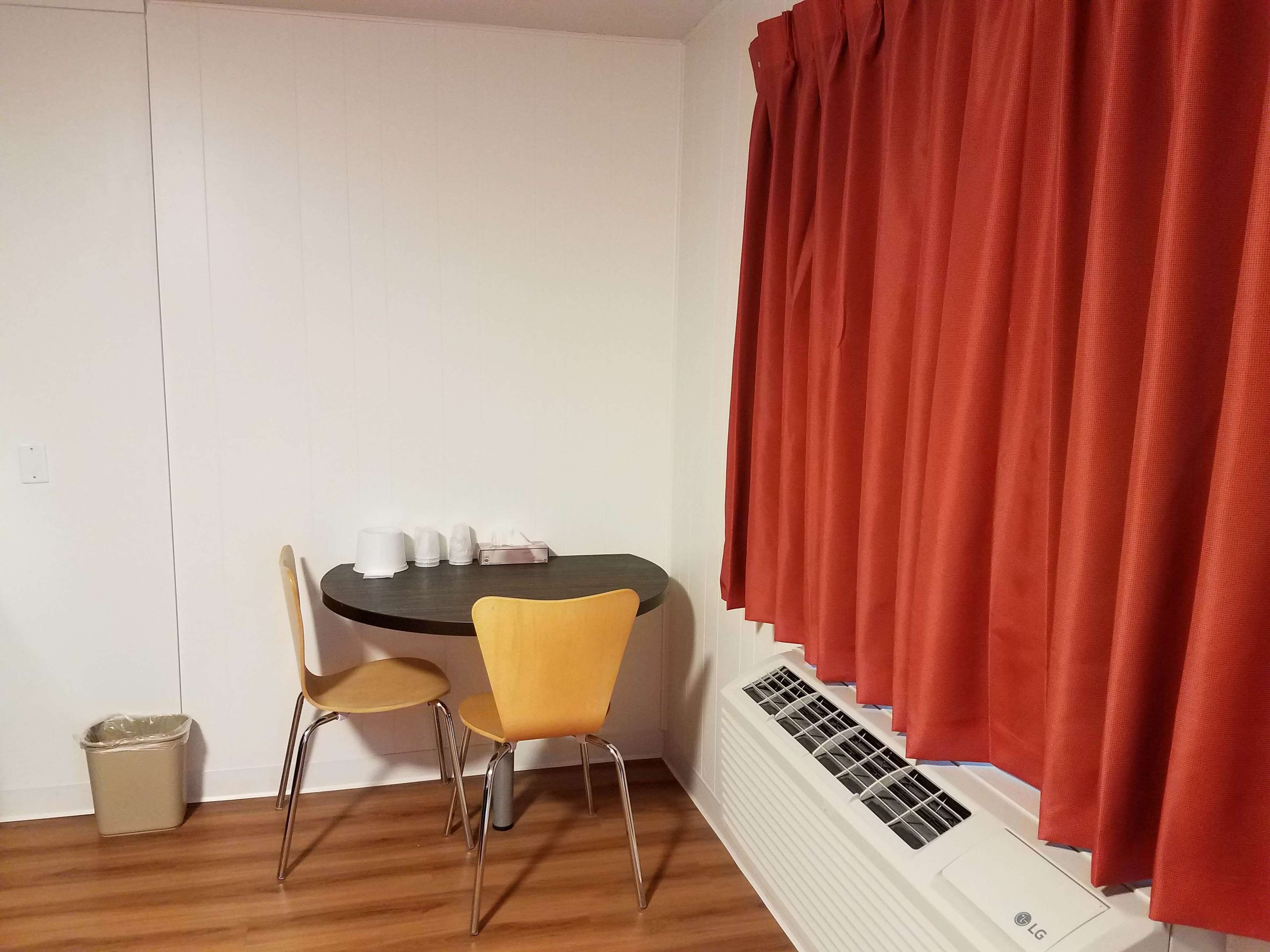 A small dining area with a round table, two chairs, a trash bin, and a window with red curtains.