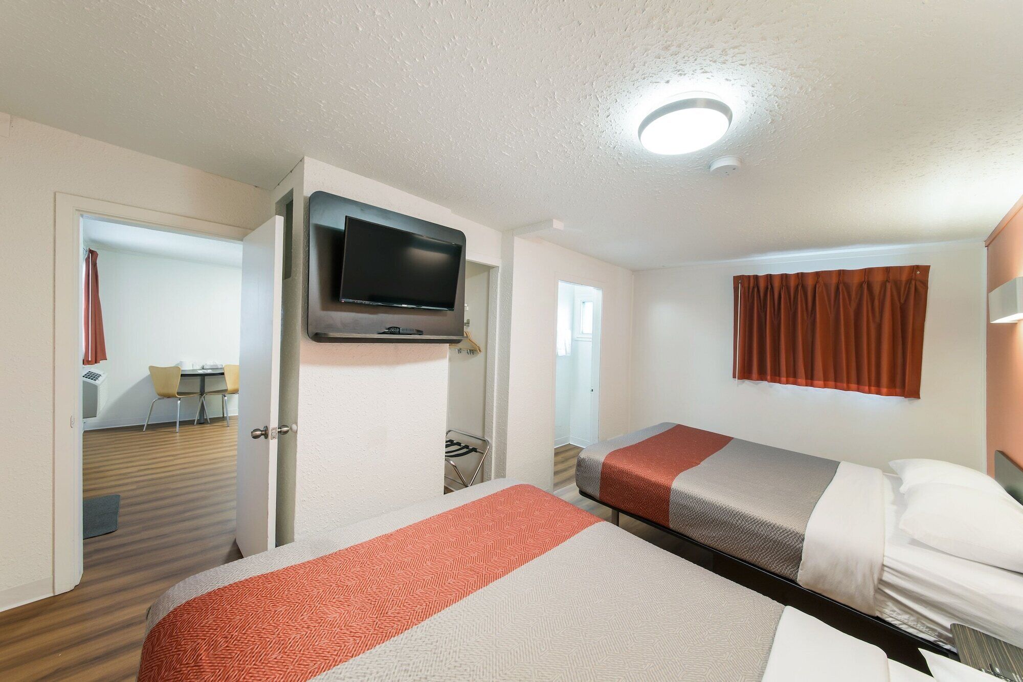 A hotel room with two beds, a television mounted on the wall, a door leading to a bathroom, and a window with curtains.