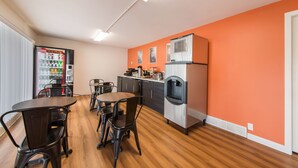 Une cuisine moderne équipée d’un réfrigérateur, d’une machine à café et d’un distributeur d’eau. On y trouve des tables et des chaises en bois, ainsi qu’une fenêtre avec des stores.