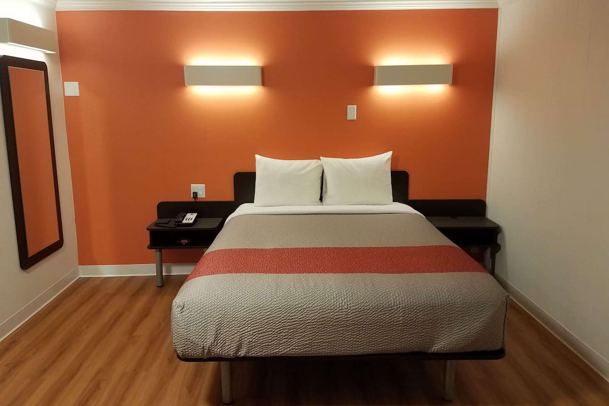 Una habitación de hotel con una pared de acento naranja, una cama con colcha a rayas blancas y rojas, dos lámparas de noche y una mesita de noche con un teléfono y un reloj digital.