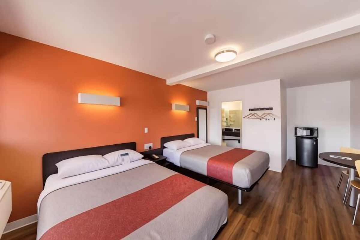Habitación de hotel con dos camas, una mesita, una silla, un cesto de basura y un espejo.
