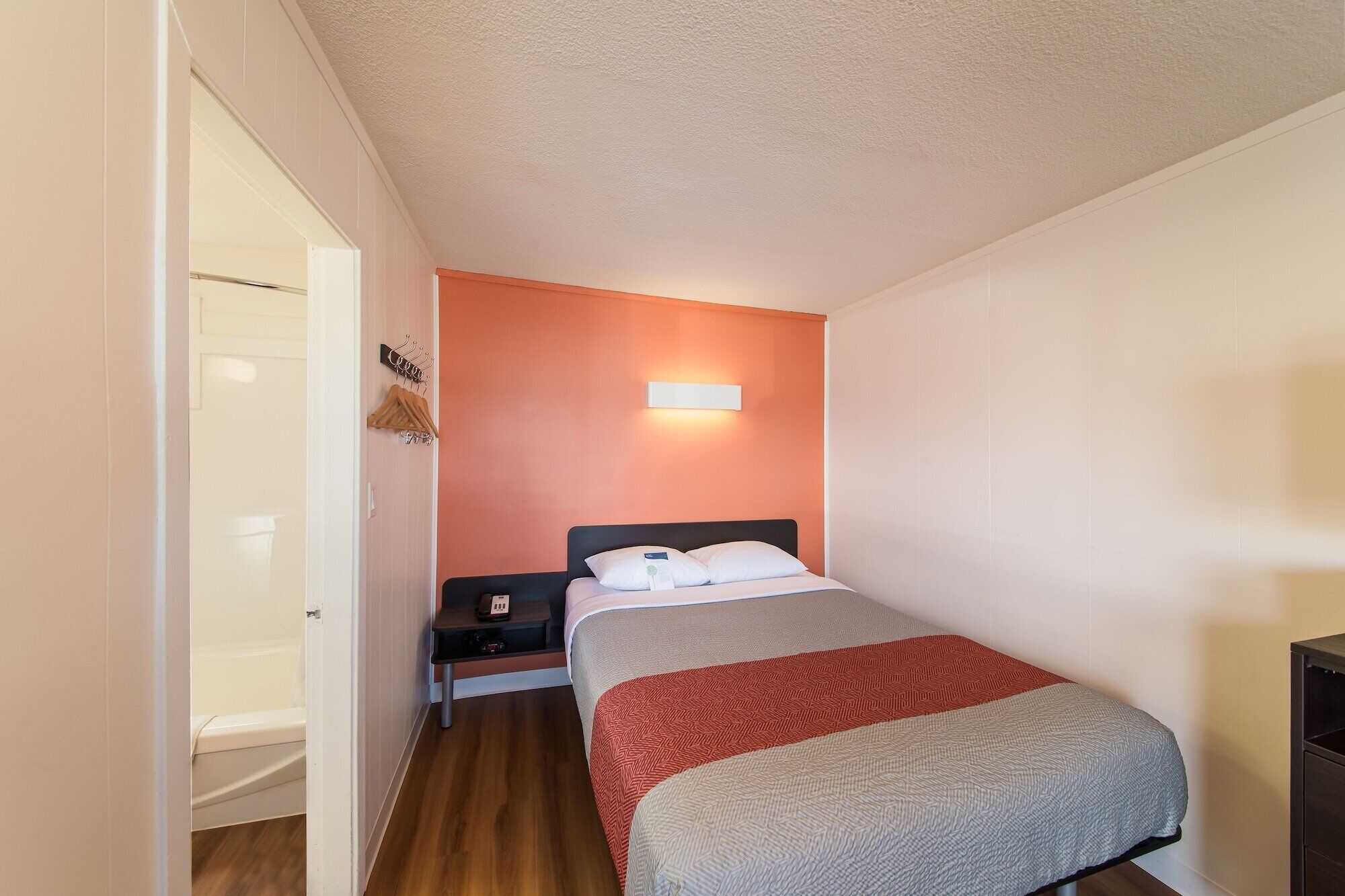 Una habitación pequeña con una cama individual, piso de madera, una mesita de noche, una lámpara fijada a la pared y una puerta que da a un baño.