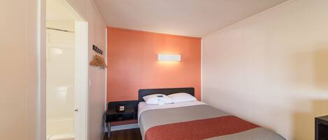 A small, single-bed room with a wooden floor, a nightstand, a bedside table, a wall-mounted light, and a door leading to a bathroom.