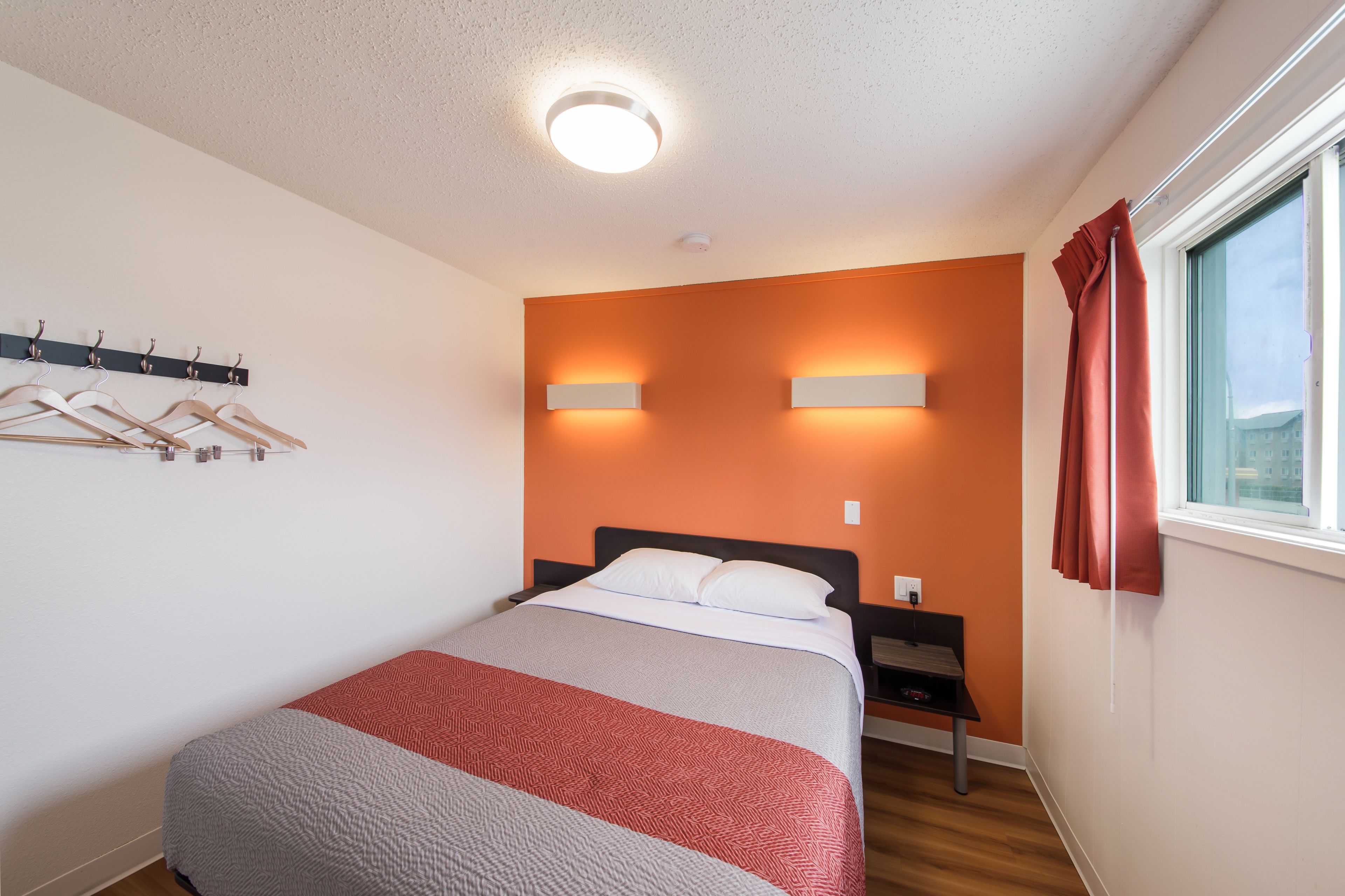 A hotel room with a bed, a nightstand, a wall-mounted light, a coat rack, and a window with curtains.