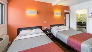 Une chambre d’hôtel avec deux lits, une fenêtre, un radiateur et une salle de bain visible à travers une porte ouverte.