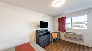 A hotel room with a bed, a desk with a flat-screen TV, a chair, a window with a view of the outside, and an air conditioning unit.
