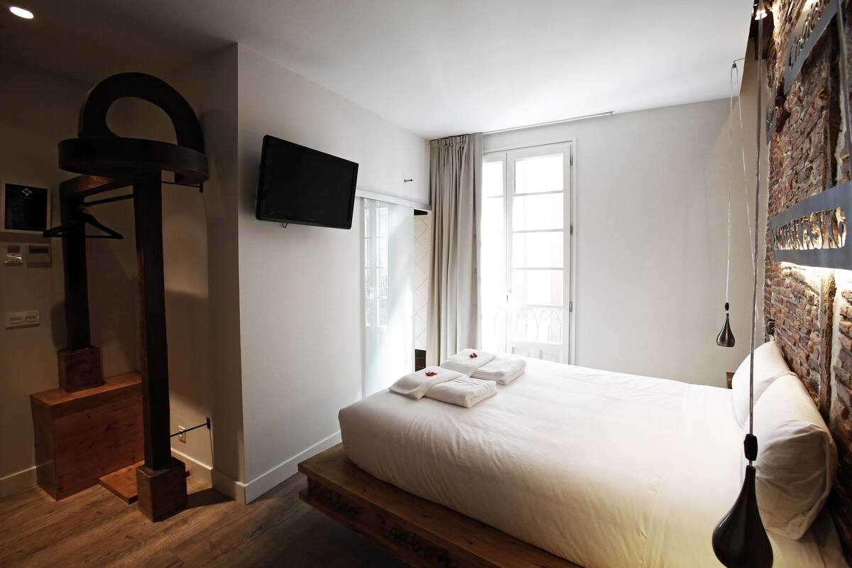 A modern hotel room with a large bed, a wooden bench, a TV, and a brick wall.
