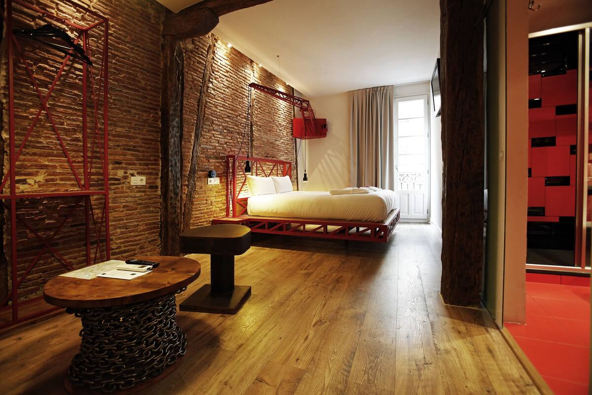 A hotel room with a brick wall, a bed with white linens, a wooden table, and a red metal ladder.