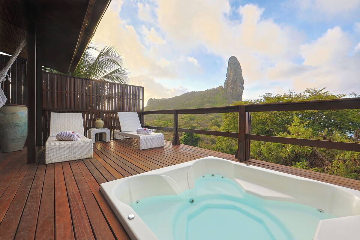 A wooden deck with a hot tub and lounge chairs, overlooking lush greenery and a prominent rock formation.