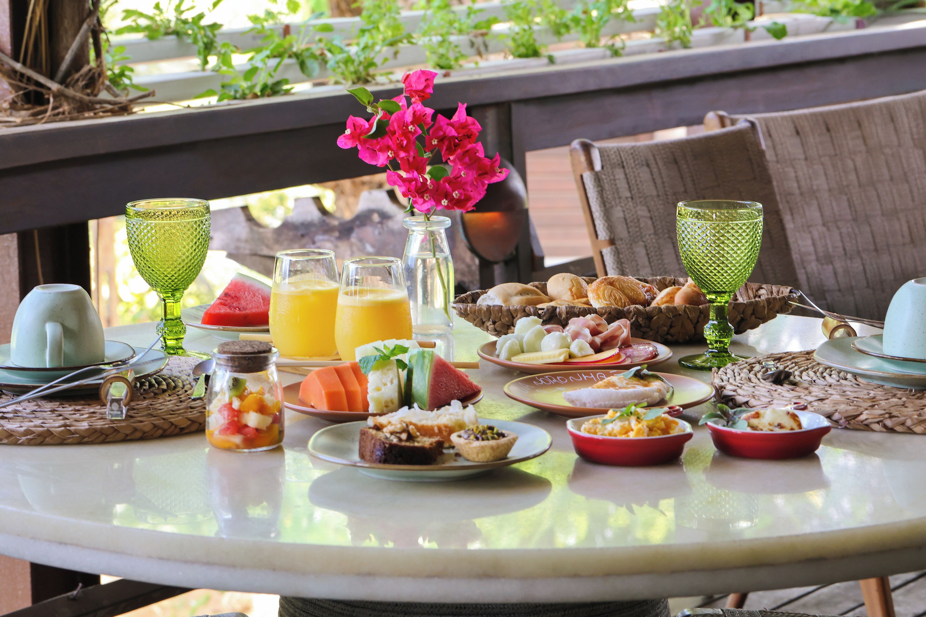 Una mesa dispuesta con diferentes alimentos para el desayuno, como frutas, bollería y bebidas, en un patio al aire libre.