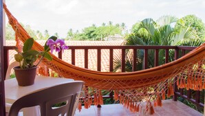 Un balcon avec un hamac, une plante en pot et une vue sur des palmiers et un toit recouvert de tuiles.
