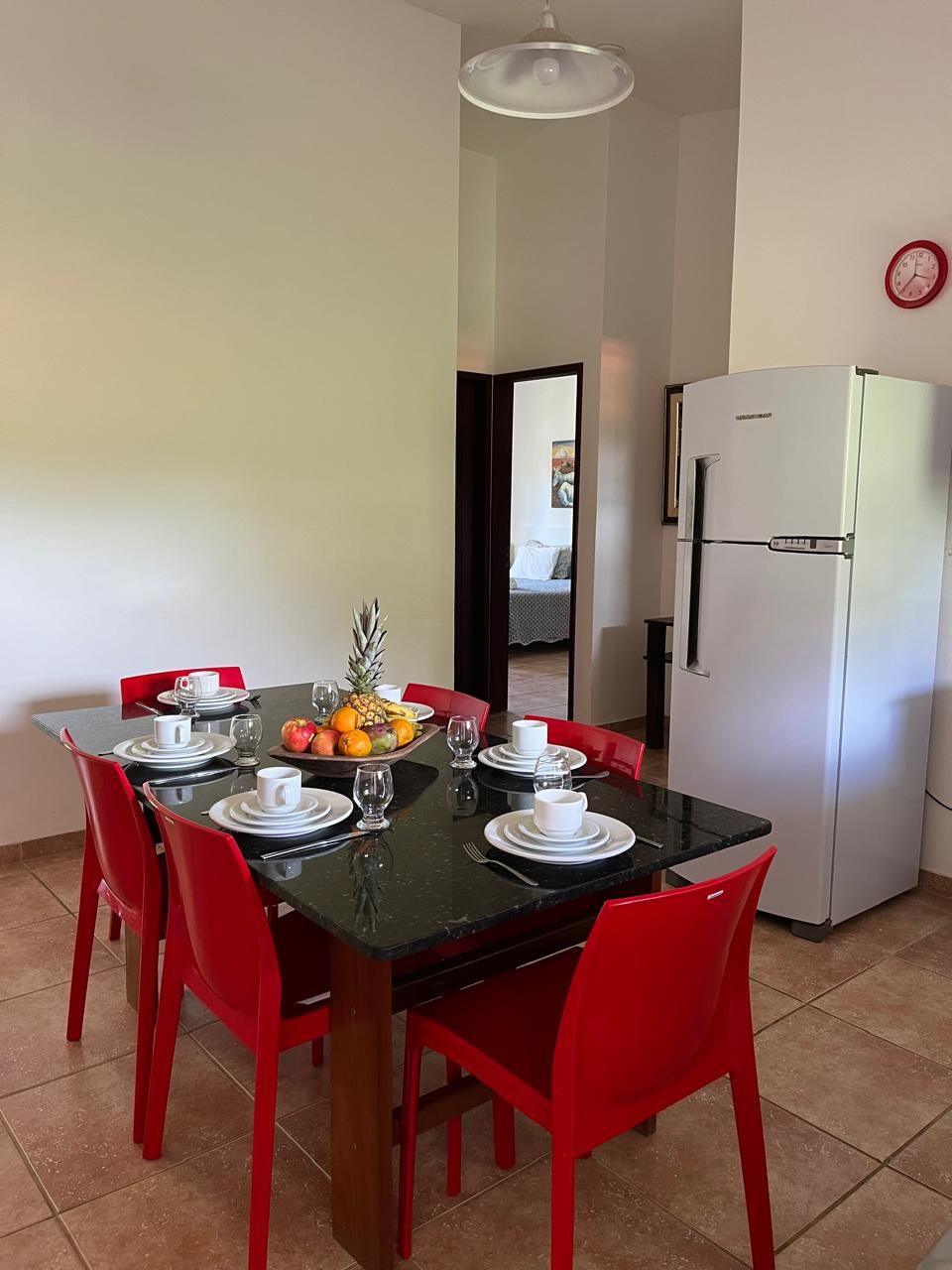 Un coin repas avec une table noire, des chaises rouges et un saladier rempli de fruits. On aperçoit un réfrigérateur blanc à l’arrière-plan.