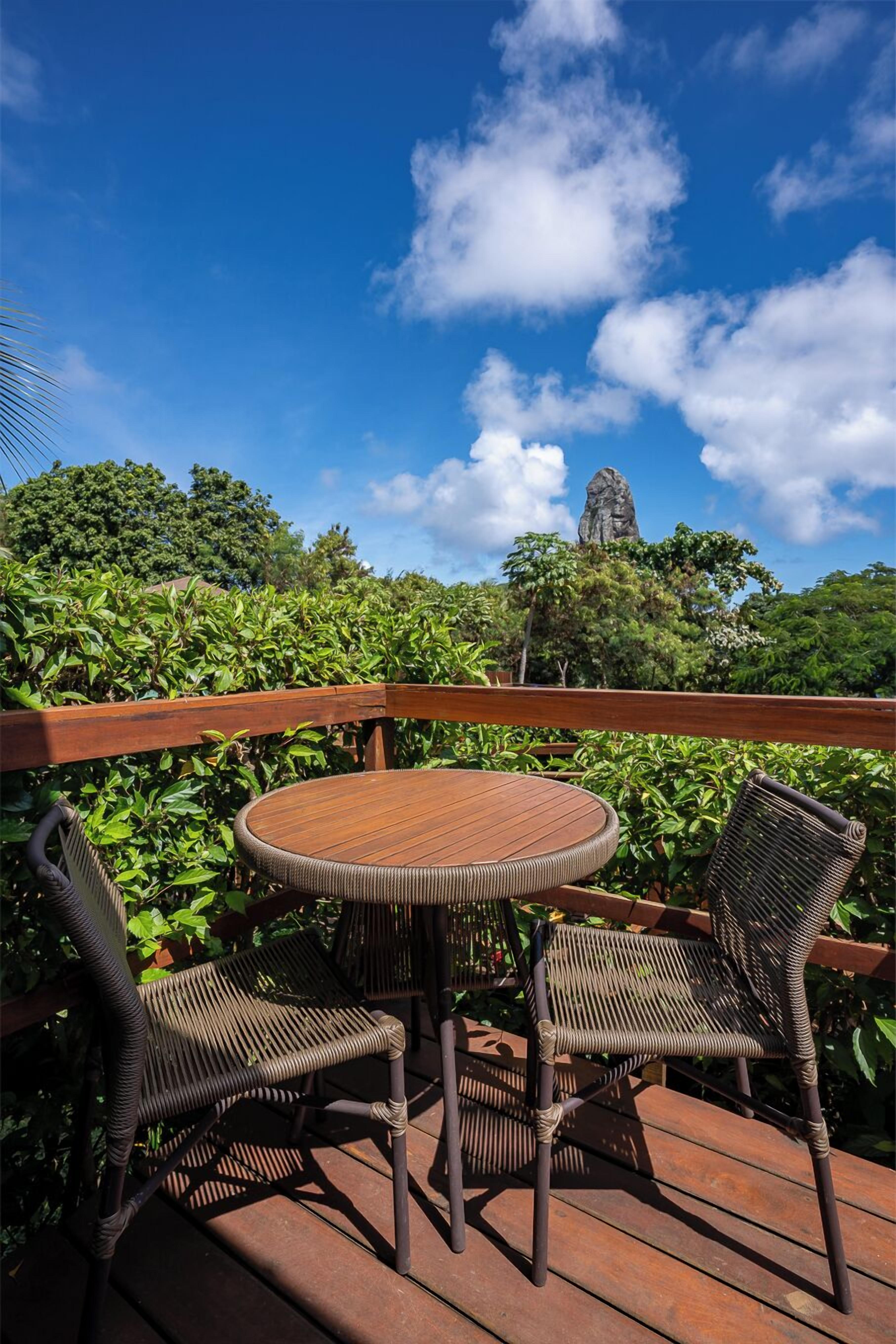 A wooden deck with a round table and two wicker chairs, surrounded by lush greenery and a clear blue sky.