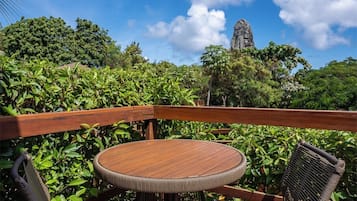 A wooden deck with a round table and two wicker chairs, surrounded by lush greenery and a clear blue sky.