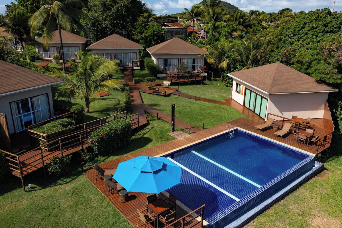 Resort com piscina, deck de madeira e vários edifícios tipo bangalô cercados por vegetação exuberante.