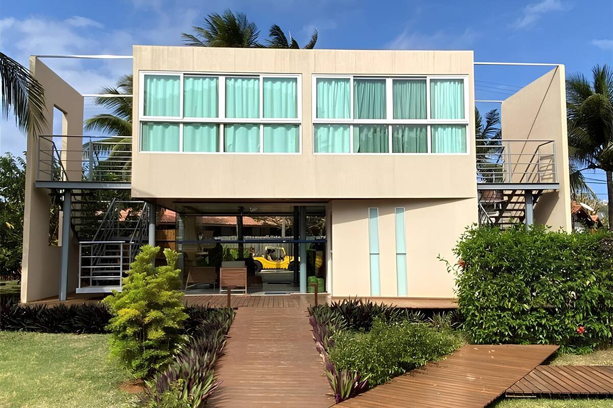 Um edifício moderno de dois andares com entrada central, área de estar coberta ao ar livre e um caminho de madeira que leva à entrada.