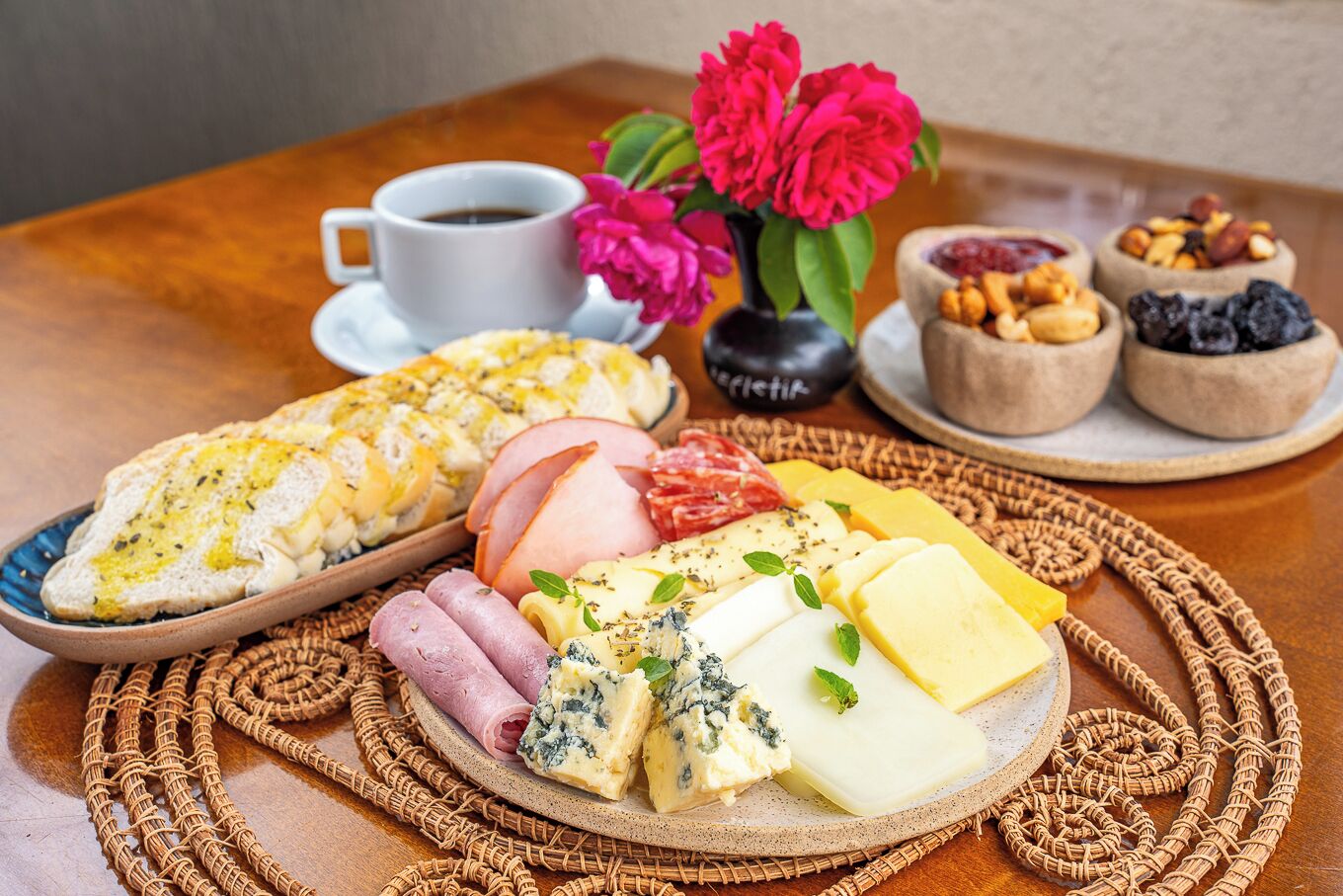 A charcuterie board with various cheeses, meats, and bread, accompanied by a cup of coffee and flowers.