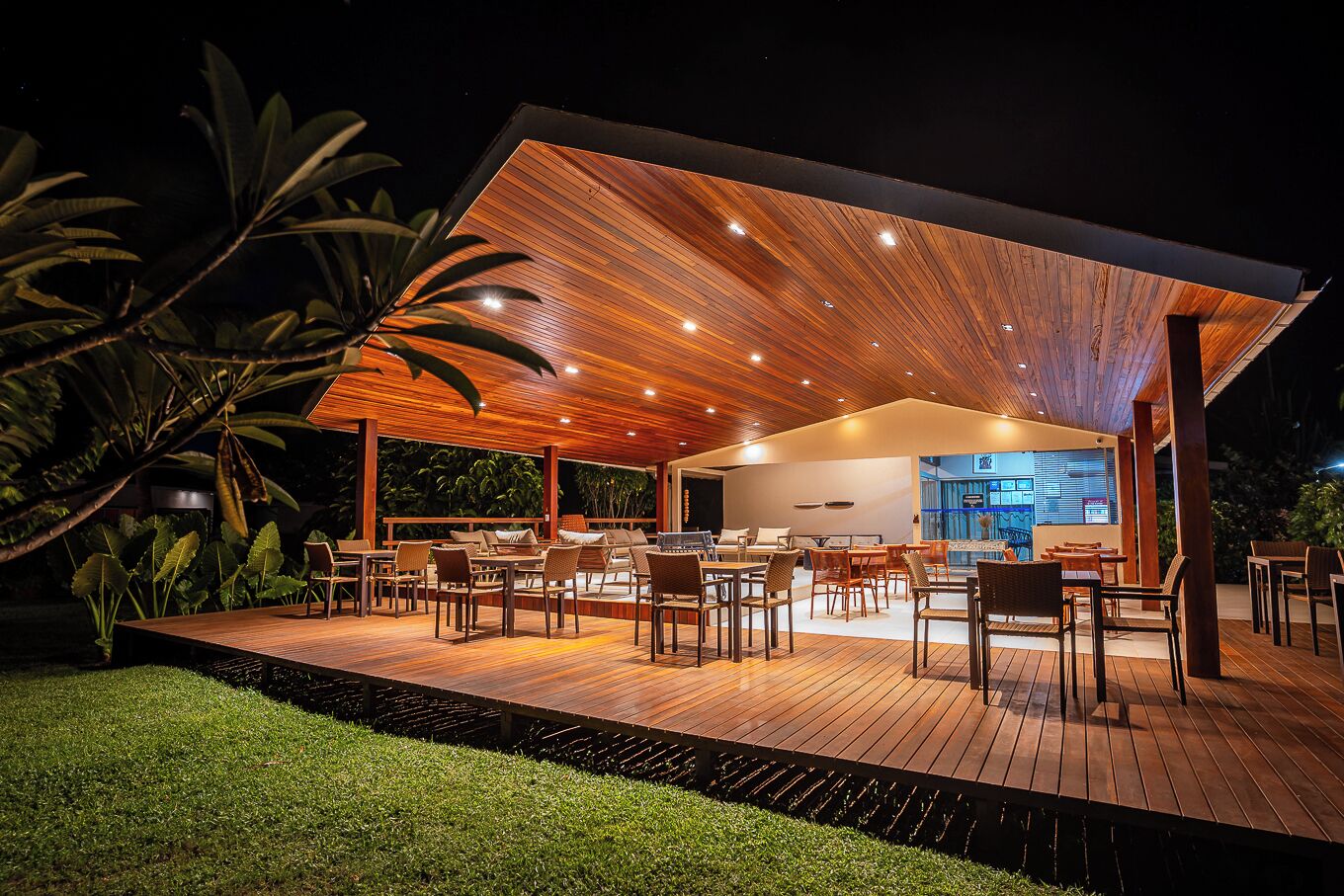 An outdoor dining area with wooden flooring and tables, surrounded by lush greenery.