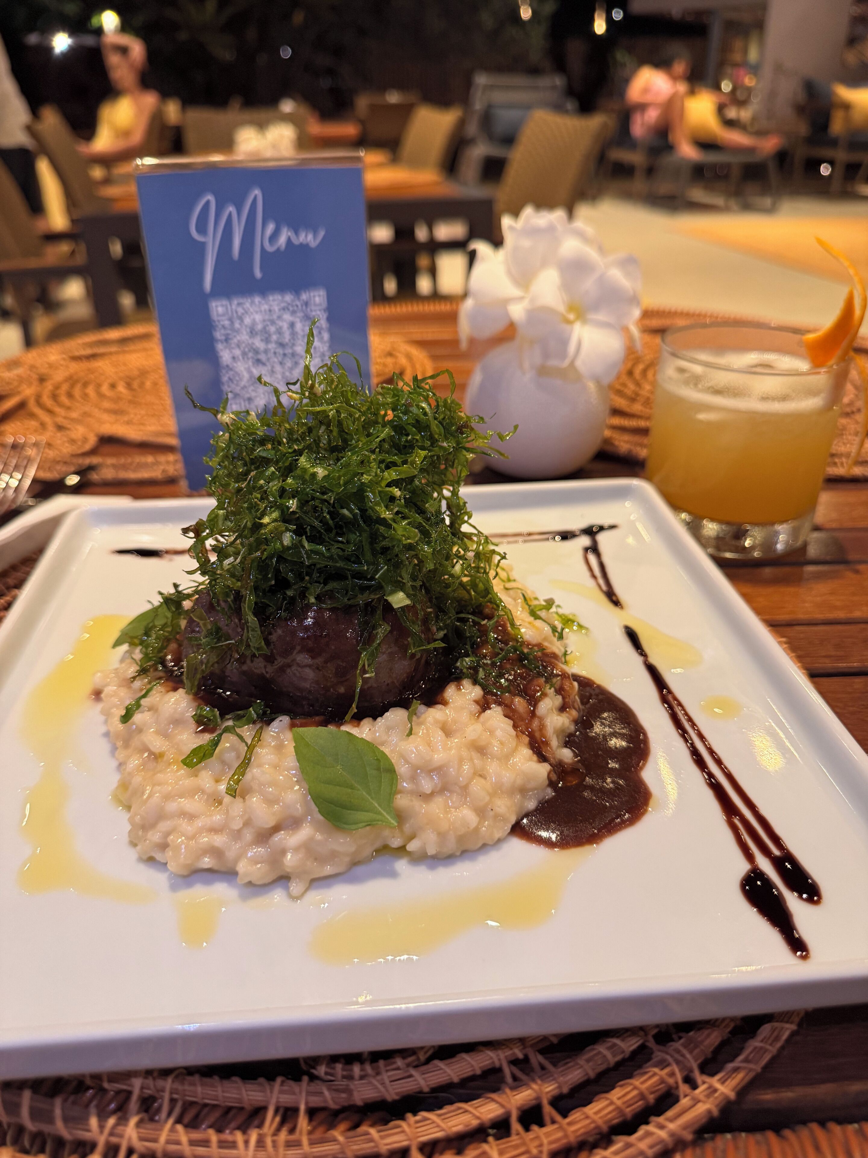 A square plate with a dish of risotto topped with greens and a dark sauce, a menu sign, and a glass of orange juice on a table.