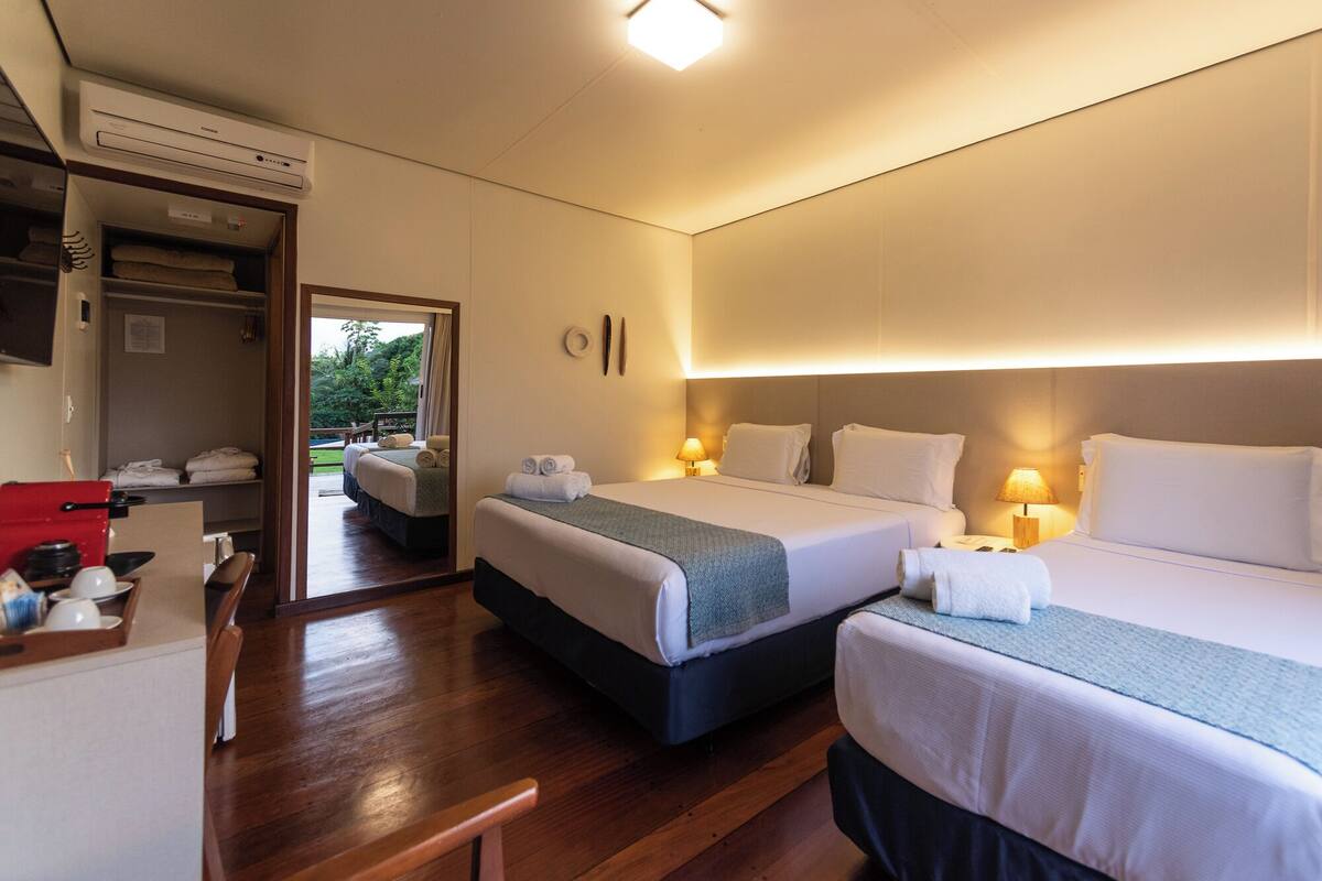 Quarto de hotel com duas camas, uma mesa e uma cadeira vermelha, e vista para o exterior.