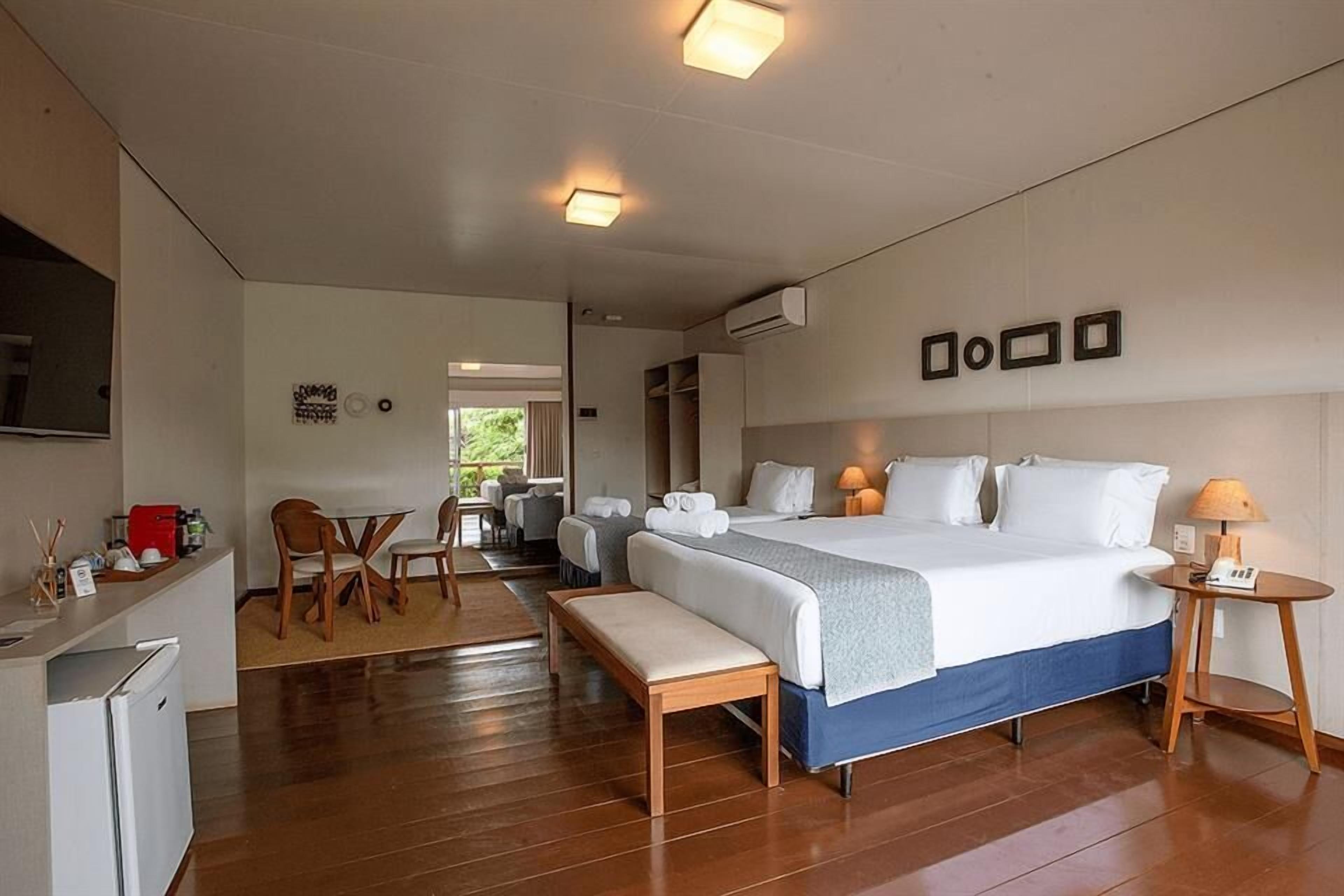 A hotel room with a large bed, a desk with a chair, a small table with a lamp, and a bench.