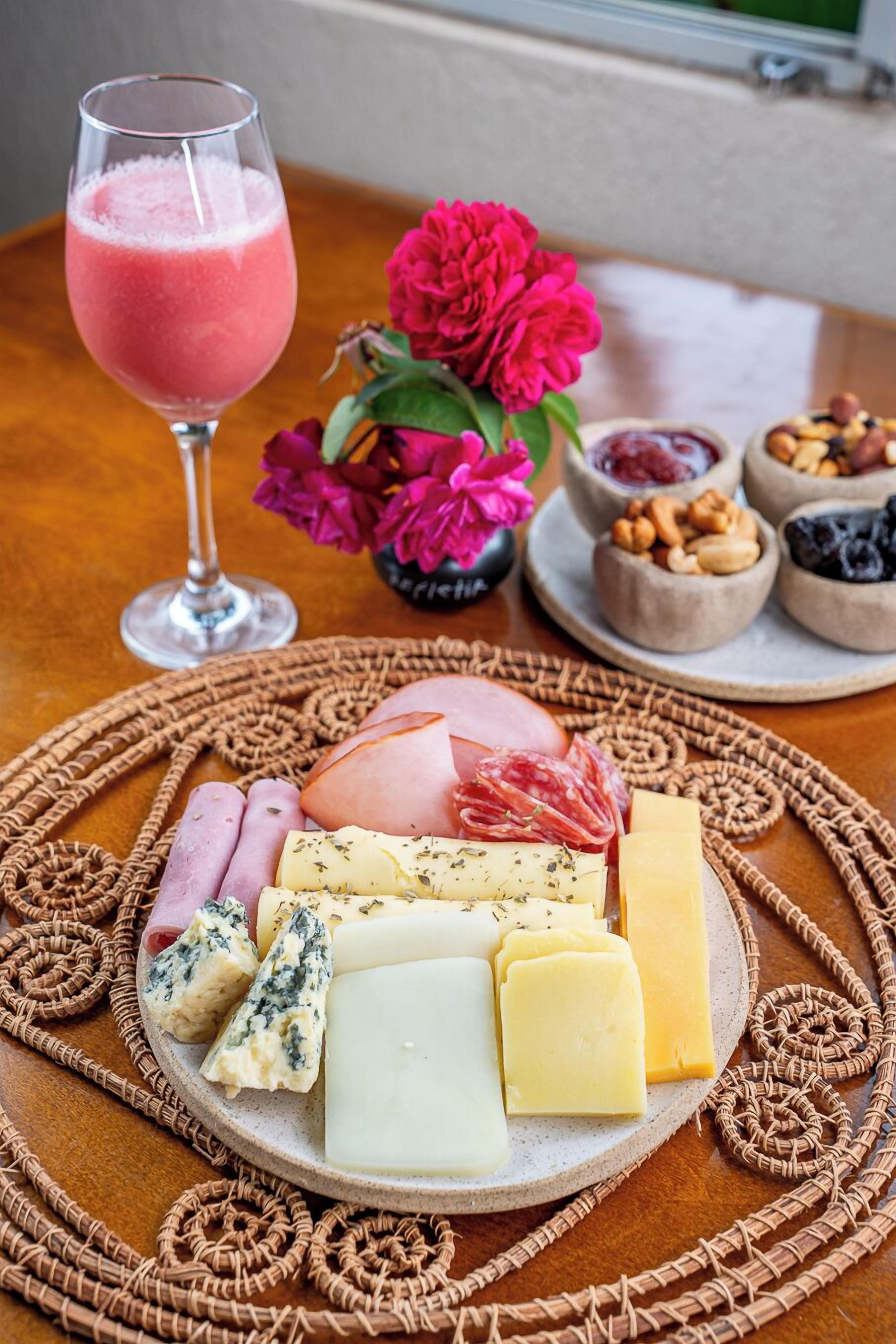 A charcuterie board with various cheeses, meats, and nuts, accompanied by a glass of pink drink and a bouquet of flowers.