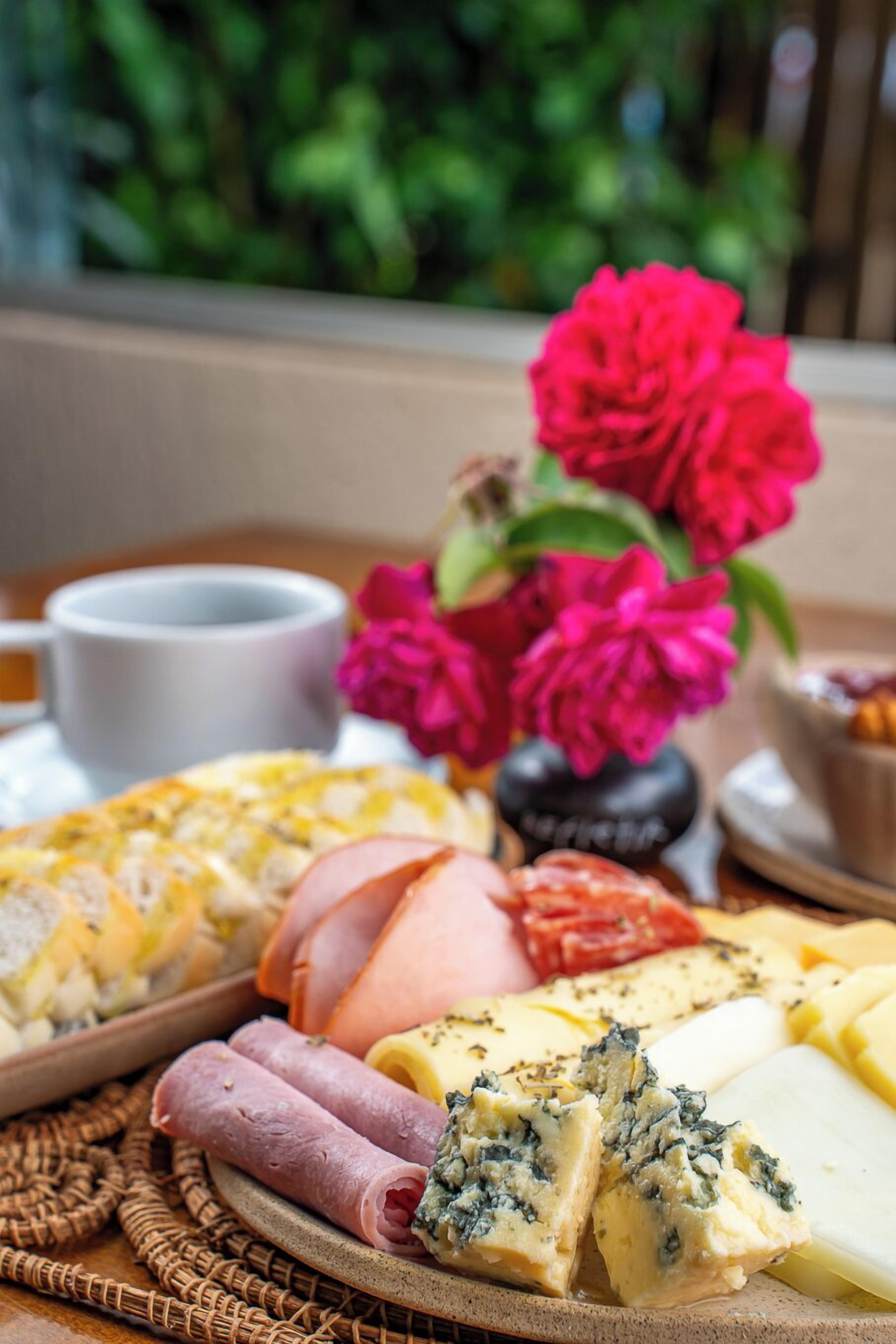 A charcuterie board with various meats, cheeses, and a cup of coffee, accompanied by a vase of flowers.
