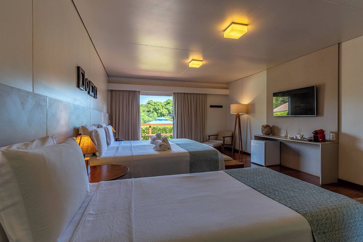Quarto de hotel com duas camas, televisão, escrivaninha e vista para o verde lá fora.