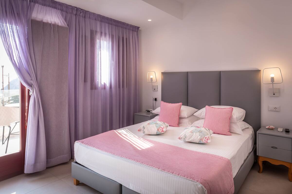 A bedroom with a large bed, two bedside tables, a nightstand, and a window with purple curtains.