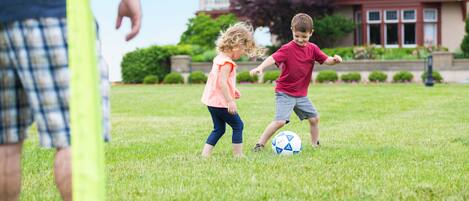 Two children playing with a soccer ball on a grassy field with a house in the background.