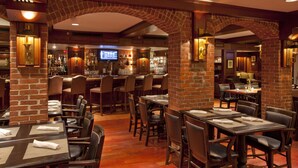 A restaurant with brick arches, wooden tables, and chairs. There is a bar area with bottles and a television screen. - The Villages at Ocean Edge (Brewster)