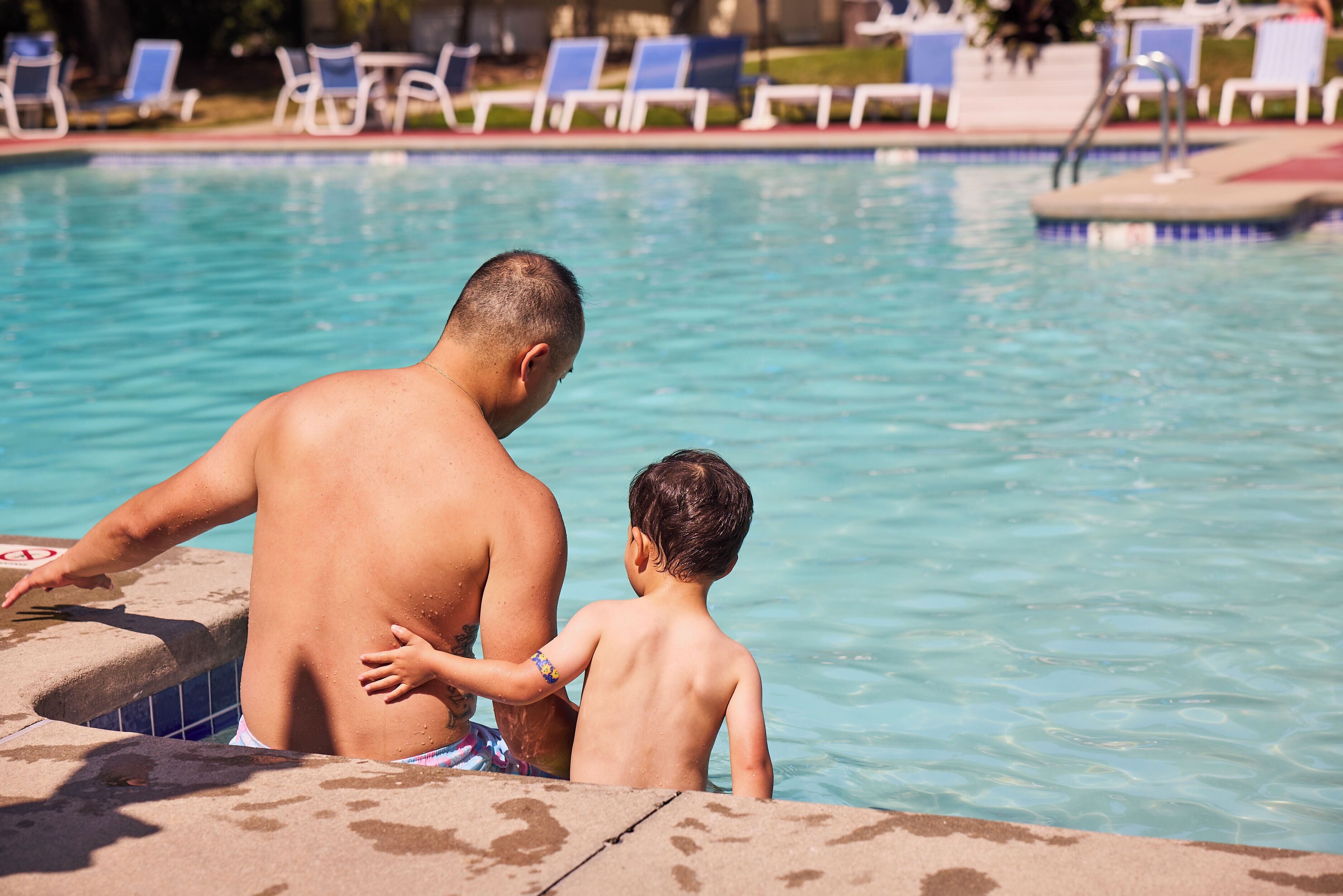 A man and a child by a swimming pool with lounge chairs in the background.