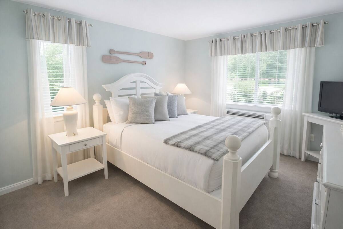 A bedroom with a bed, nightstands, a TV, and windows with curtains.