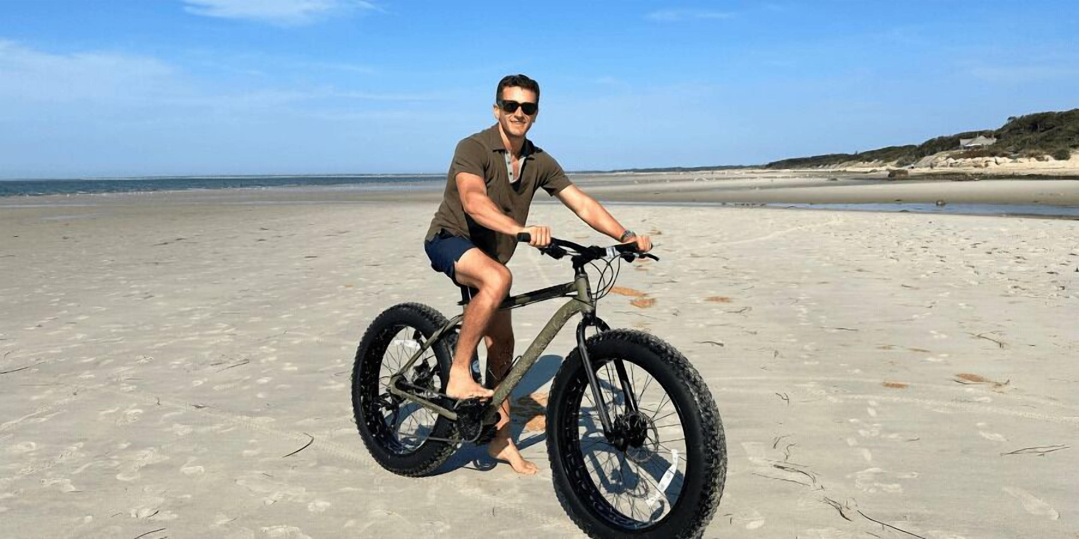 A person riding a bicycle on a sandy beach with the ocean in the background.