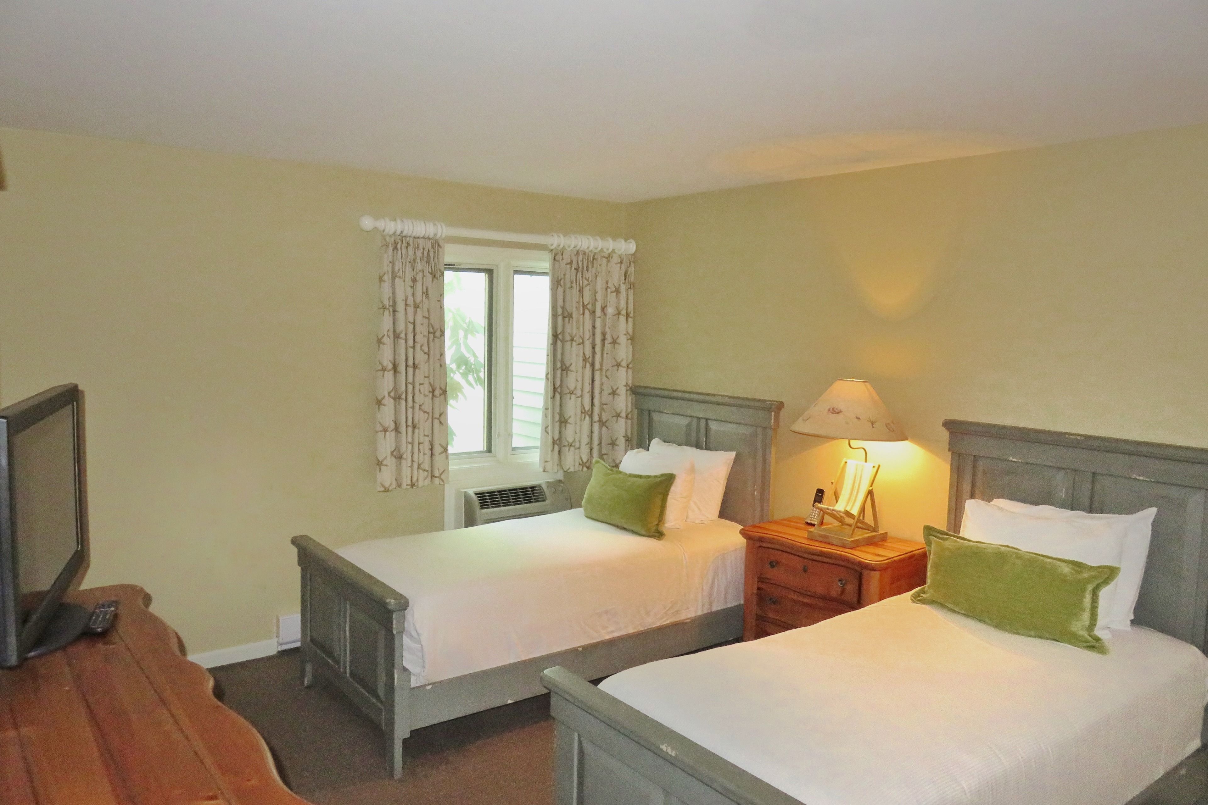 A room with two single beds, a bedside table with a lamp, a TV, and a window with curtains.