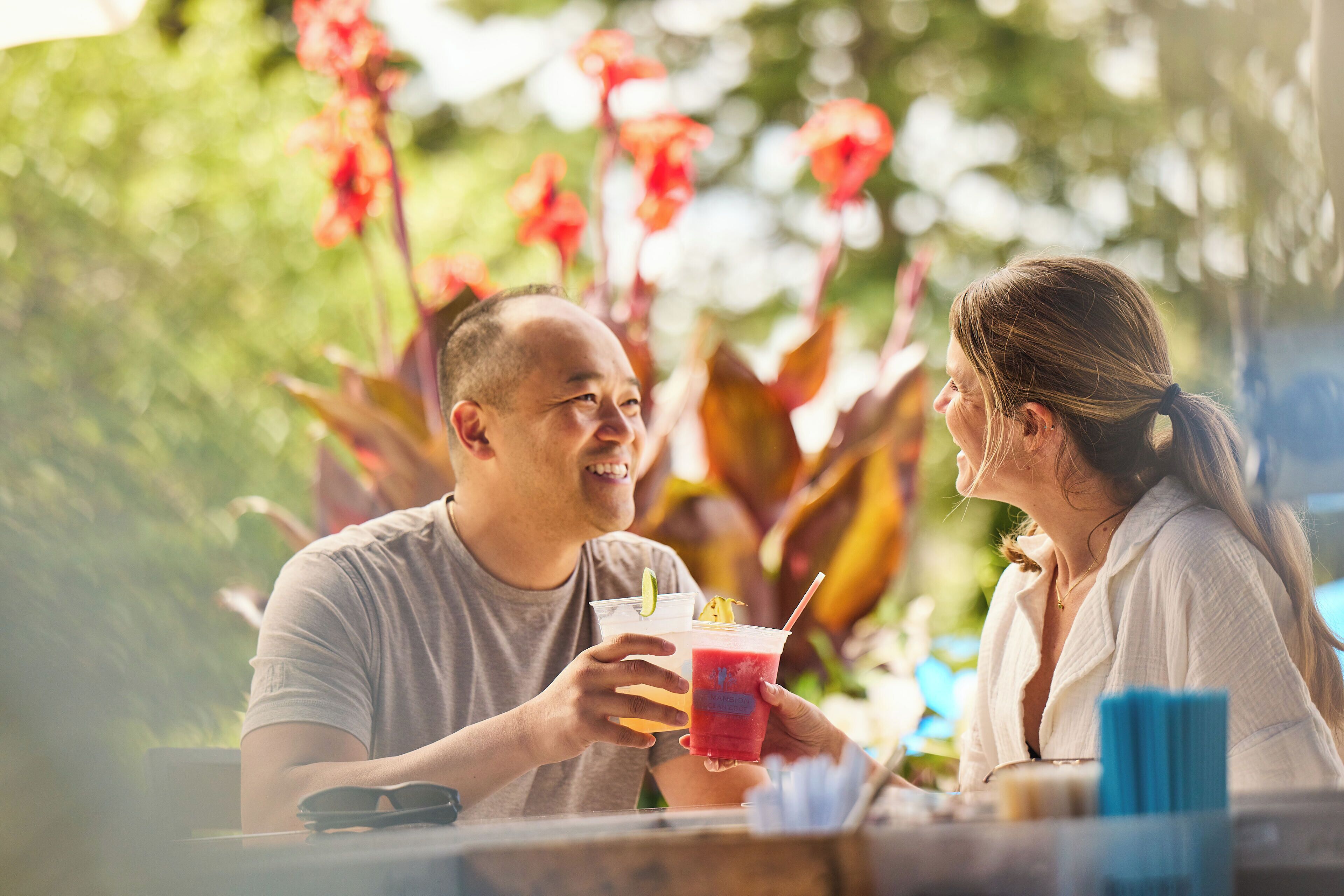 Two people sitting at a table with drinks, surrounded by greenery and red flowers.