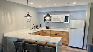 A modern kitchen with a large island, wooden cabinets, a white refrigerator, and pendant lighting. - The Villages at Ocean Edge (Brewster)