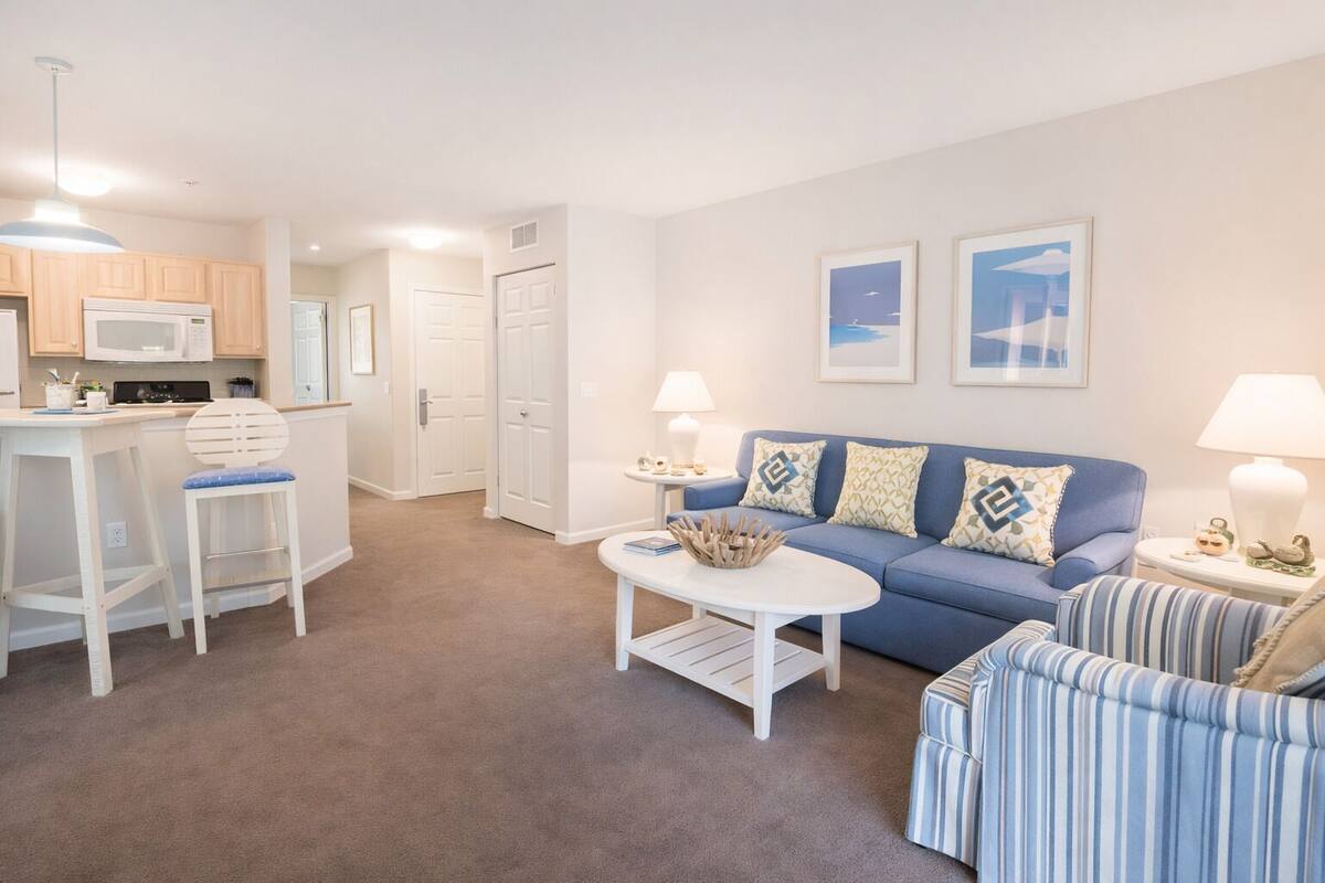 A living room with a blue sofa, striped armchair, and a white coffee table. There is a kitchen area with a microwave and a bar stool.